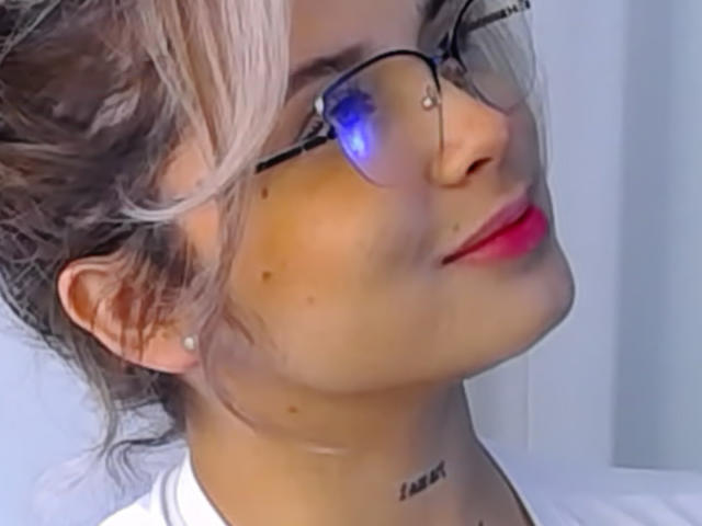KateZoe mit Brille, rosafarbenen Lippen, blonden Haaren und einem kleinen Muttermal auf ihrer Wange, hat ein Tattoo unter ihrem Kinn und einen blauen Lichtreflex in ihren Augen..