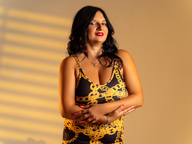 MadameAlexaX with curvy body, black and yellow patterned dress, large boobs, black wavy hair, red lipstick, hands crossed, gold necklace, and bracelet, standing in soft light.