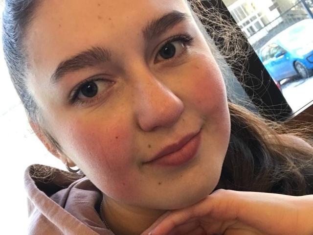 Anabellis with a slight smile, brown eyes, and pinkish cheeks, has her hand under her chin. She has a small mole under her right eye and brown hair.
