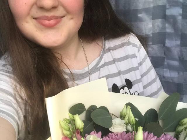 Anabellis with light pink lips and brown hair, wearing a gray-striped shirt, holding a bouquet of pink and white flowers with green eucalyptus leaves.