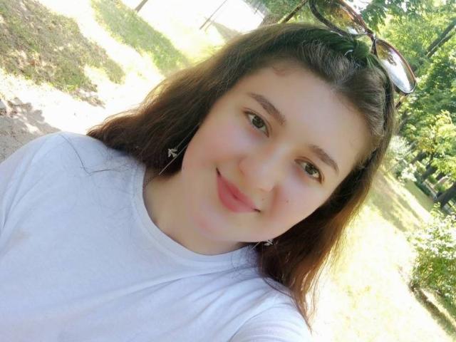 Anabellis with pale skin, brown eyes, and long brown hair, wearing a white shirt, smiling softly, has small earrings, and sunglasses on her head, in a sunny outdoor setting.
