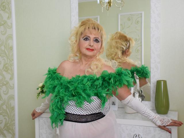 MarthaBrownn with curly blonde hair, wearing a green feather off-shoulder top, white polka-dot dress, and white gloves, standing in front of a mirror, smiling. Boobs visible.