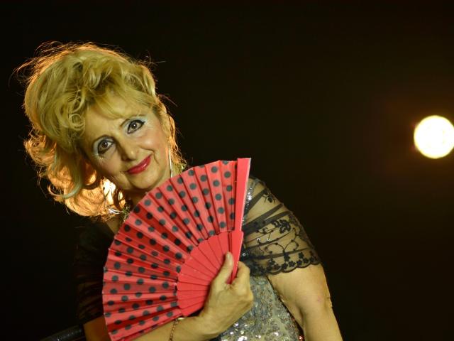 MarthaBrownn with curly blonde hair, holding a red fan with black dots, wearing a black lace top, and a shiny silver dress, smiling at the camera.