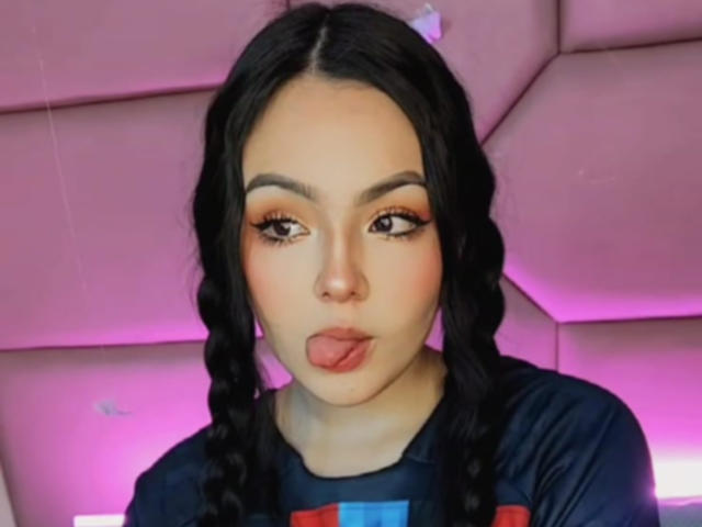 HaleiBeckham met volgeblazen lippen, tong uitgestoken, gevlochten zwart haar, oranje oogschaduw, donkere wenkbrauwen, draagt een zwarte top, achtergrond met roze gloed, zachte belichting, schattig, sexy, vrouwelijk..