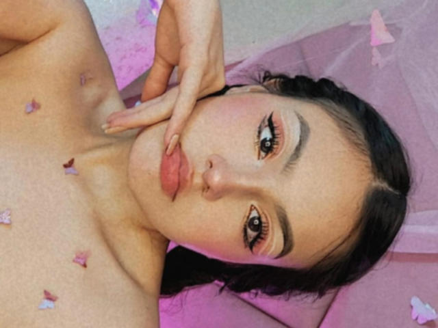 HaleiBeckham, avec ses lèvres pincées, ses cheveux foncés et son fard à paupières rose, touchant son visage, avec des pétales de fleurs en forme de papillons éparpillés sur sa poitrine nue, regardant l'appareil photo..