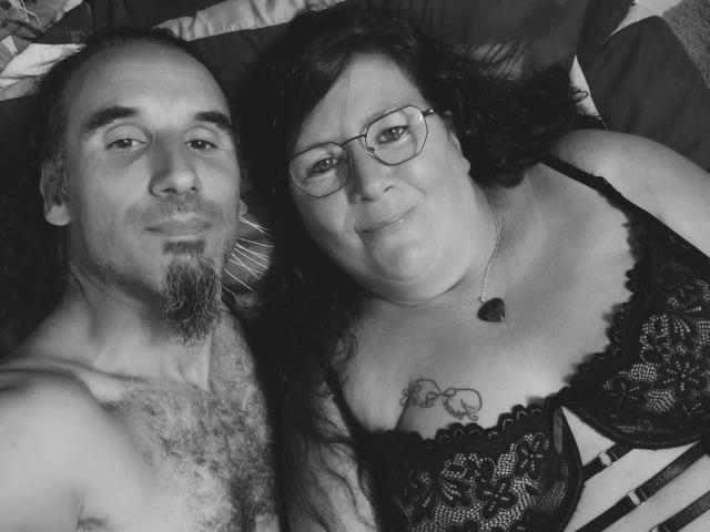 InstantShow69 com barba e pelos no peito, sem camisa, deitado ao lado de InstantShow69 usando um sutiã de renda preto, óculos, colar de coração e tatuagem no peito, em uma selfie em preto e branco..