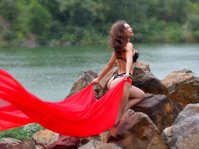 AurikaBriz, con cabello castaño, lencería negra y tela roja que fluye, sentada en una orilla rocosa, sosteniendo un paño rojo, con los ojos cerrados, de espaldas a la cámara, mostrando su trasero..