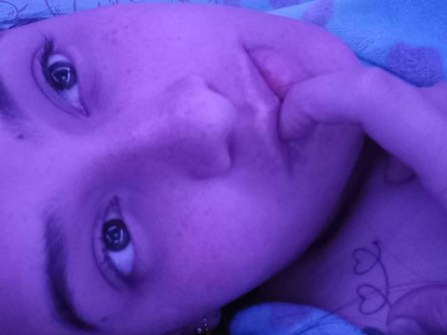 VictoriaF69's face in purple light, freckled, with her finger on her chin, looking up, visible tattoo on her neck, blue patterned blanket, slightly parted lips.