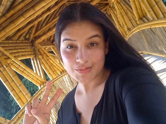 VictoriaFer69 with long dark hair, light skin, and brown eyes, wearing a black zip-up shirt, making a peace sign, smiling under a yellow bamboo roof.