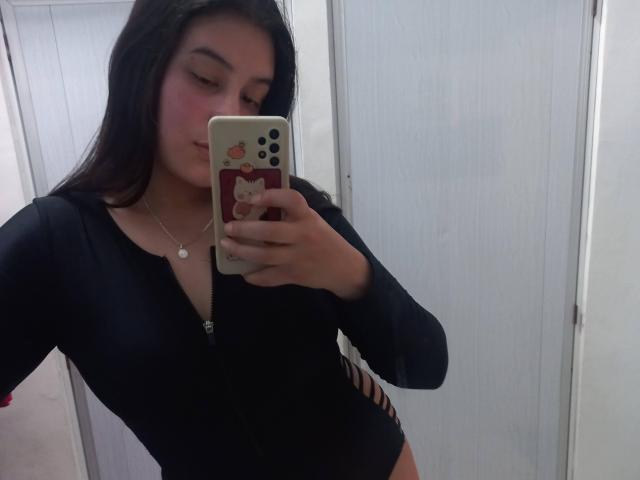 VictoriaFer69, com cabelos longos e escuros, vestindo um macacão preto com zíper, segurando um celular com uma capa de gato, mostrando suas nádegas através de recortes nas laterais, em uma selfie no espelho..