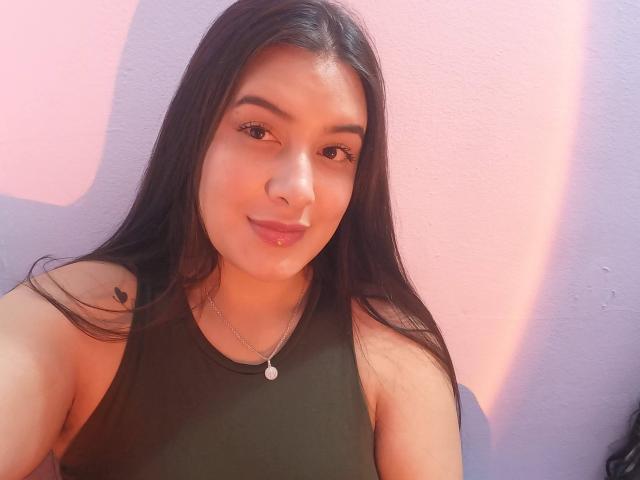VictoriaFer69, met lang zwart haar, bruine ogen en een schattige glimlach, draagt een groene tanktop, een kleine ketting en een kleine tatoeage op haar rechter schouder. Ze is een sexy Latina..