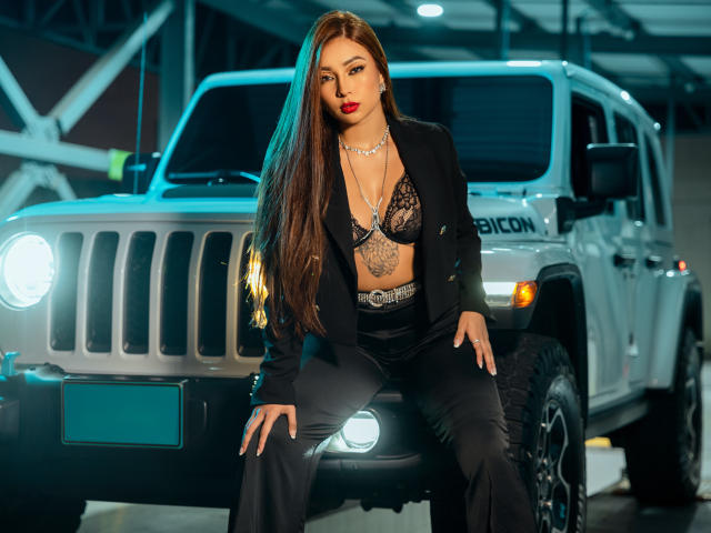 IrisStone, con lunghi capelli castani, rossetto rosso, reggiseno di pizzo nero e un blazer, seduta su un Jeep bianco, che ostenta il seno e i pantaloni neri in un garage..