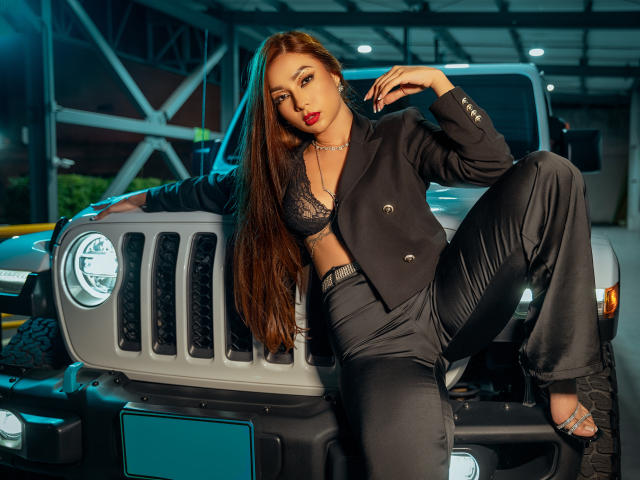 IrisStone leunt tegen een grijze Jeep, gekleed in een zwart blazer, leren broek en een zwarte kanten BH, met lang bruin haar en rode lippenstift, waarbij haar borsten en billen zichtbaar zijn..