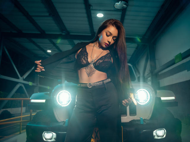 IrisStone with long brown hair, black lace bra, and high-waisted pants, pulling a black jacket, standing in front of a car with bright headlights, showing her tits.