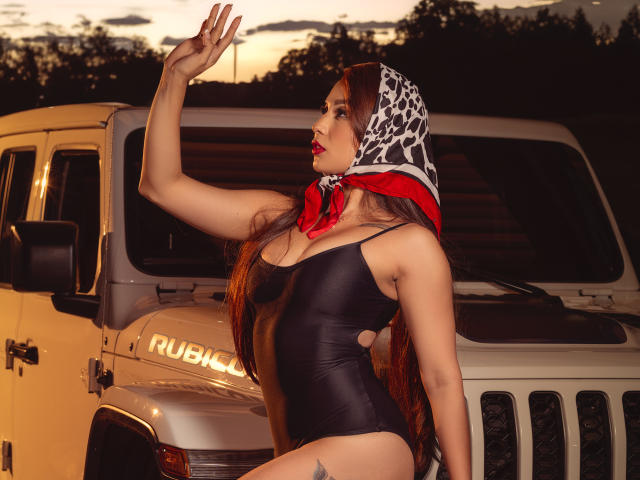 IrisStone, con lunghi capelli castani, sciarpa a stampa leopardo, bandana rossa, costume intero nero lucido, appoggiata a un Jeep bianco, con un braccio alzato, al tramonto, con un tatuaggio sulla coscia sinistra, sicura di sé, sexy..