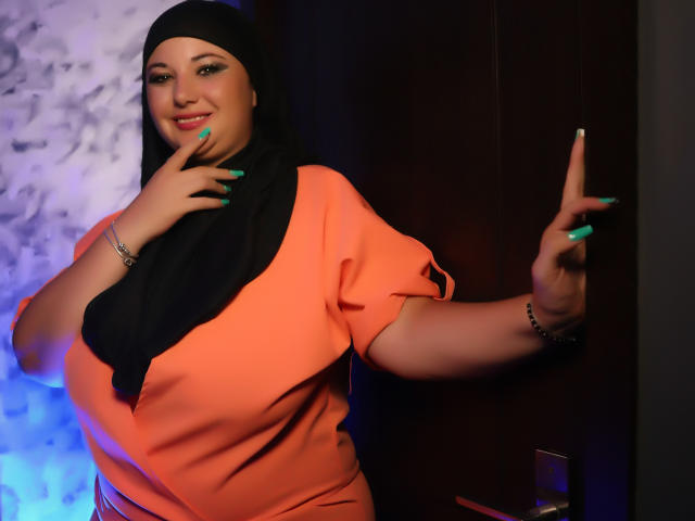AleshaArabian, com hijab preto, vestido laranja, unhas verdes, tocando a bochecha, mão direita na parede escura, sorrindo, pulseira de prata, em pé, porte médio, fofa, sexy, pele escura..
