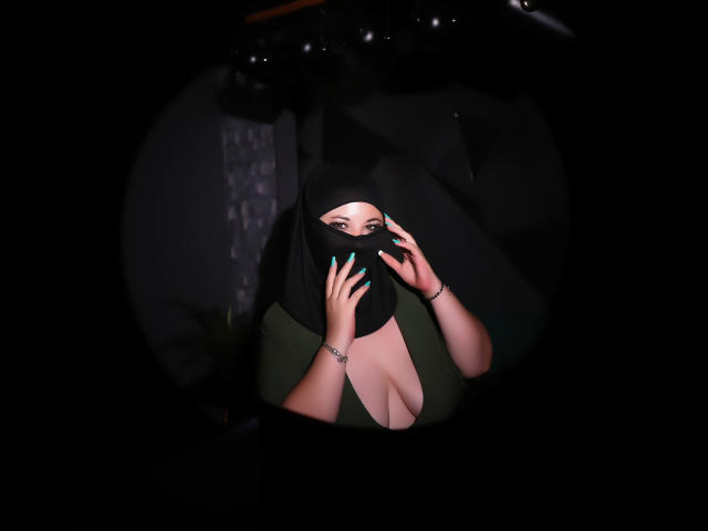 AleshaArabian with medium tits, wearing a black mask and green dress, touches her face with teal nails, visible in a dark, circular frame.
