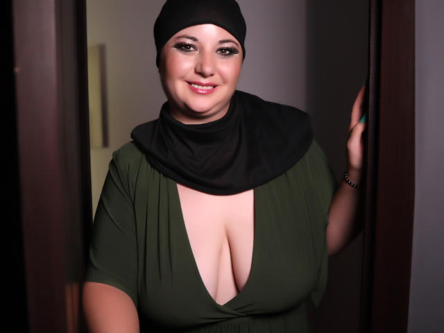 AleshaArabian with big tits, green dress, deep cleavage, black headscarf, smiling, holding door frame, right hand, dark brown eyes, light skin, pink lipstick.