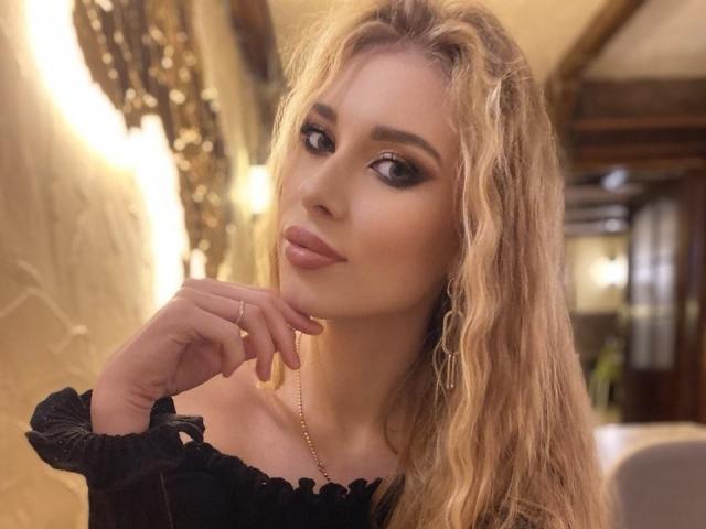 MaudieMel, con cabello rubio ondulado, ojos marrones seductores y labios carnosos, vestida con una blusa negra con un hombro descubierto, se toca la barbilla con un collar dorado que se ve..
