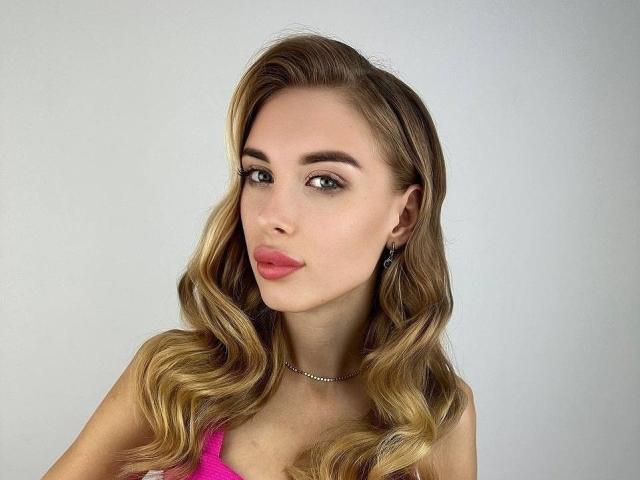 MaudieMel tiene el cabello rubio y ondulado, ojos verdes, lápiz labial rosa y un collar de plata. Lleva una blusa rosa y tiene una expresión ligeramente fruncida, sexy..