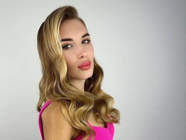 MaudieMel has blonde wavy hair, pink lipstick, and green eyes. She's wearing a pink top and looking over her shoulder with a slight smile. Her tits are partially visible.