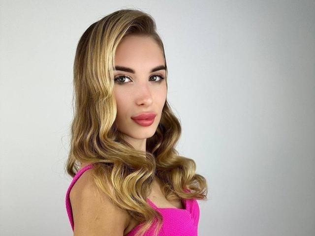 MaudieMel heeft blond, golvend haar, roze lippenstift en groene ogen, draagt een roze top en kijkt met een lichte glimlach in de camera, waarbij haar schattige achterkant zichtbaar is..