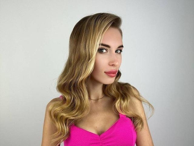 MaudieMel, con cabello rubio ondulado, una blusa rosa y un maquillaje discreto, mirando a la cámara con una ligera sonrisa, lleva un collar delgado y muestra sus pequeños senos..