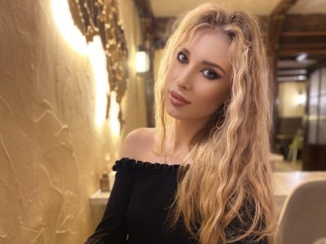 MaudieMel tem cabelos loiros ondulados, um rosto bonito e seios pequenos. Ela está vestindo uma blusa preta com um ombro de fora, com maquiagem discreta e um leve sorriso na foto..