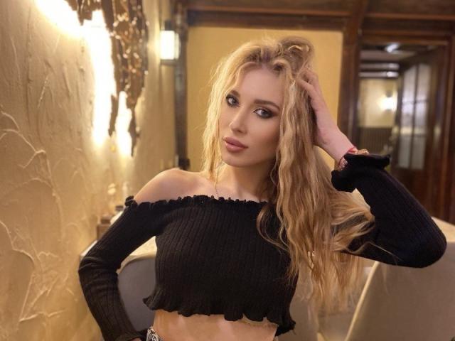 MaudieMel met golvend blond haar, die een zwarte crop top met open schouder draagt, met haar hand in haar haar, die iets opzij kijkt, in een warm verlichte binnenomgeving..