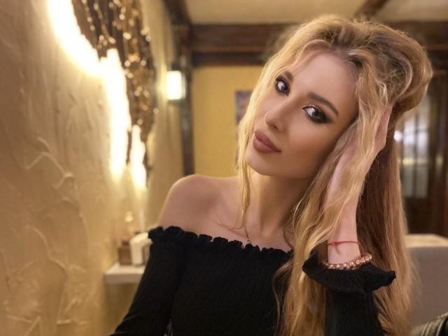 MaudieMel con cabello rubio, blusa negra con un hombro descubierto, mano en el cabello, maquillaje sutil, sonrisa suave, luz tenue, ojos marrones cálidos, labios ligeramente fruncidos, labial discreto, ambiente interior acogedor..