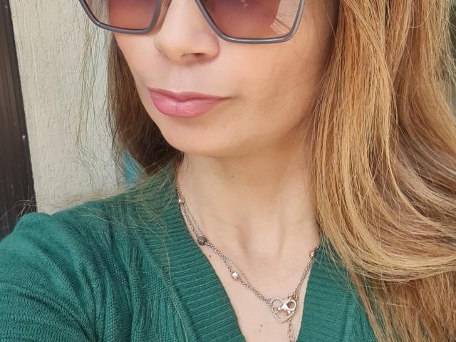 GiovannaSweet tem pele clara, lábios rosados e usa óculos de sol marrons grandes. Ela veste uma camiseta verde texturizada com um colar de coração e tem cabelo loiro e ondulado..