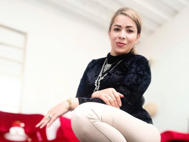 DalilaSuperHot with blonde hair, black patterned top, white pants, gold necklace, and red lipstick, sitting and smiling, showing her ass slightly. Blurred white ceiling in background.