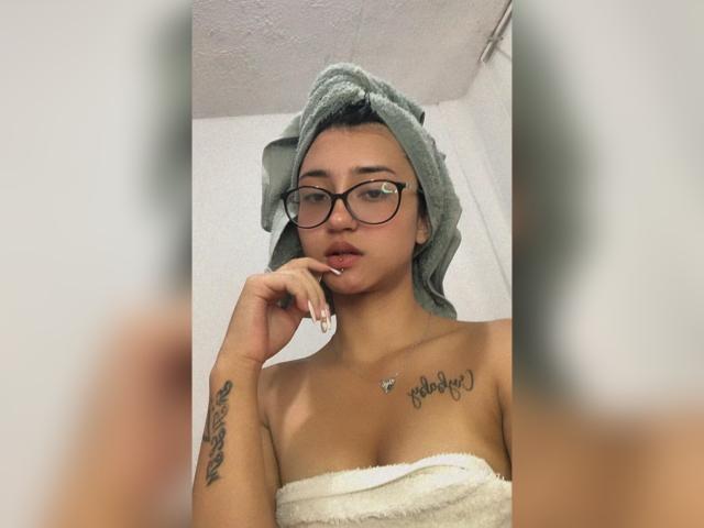 Lucia Calixto, con una toalla en la cabeza, gafas y un piercing en el labio, tocándose los labios, mostrando sus pechos, un tatuaje en el brazo izquierdo y otro en el pecho..