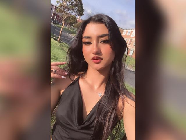 LuciaCalixto with long black hair, wearing a black halter dress, pouty red lips, and a necklace, standing outdoors, hands near face, soft sunlight, blurred greenery.