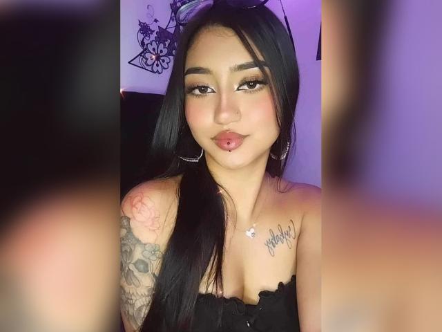 Lucia Calixto, met lang zwart haar, oorringen en een zwart topje zonder bandjes, toont een tatoeage op haar rechter schouder en een lip piercing, en kijkt naar de camera..