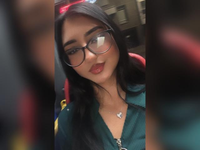 Lucia Calixto, con cabello negro largo, gafas, labios rojos y una blusa verde, mostrando un pequeño piercing en la nariz, lleva un collar y está sonriendo ligeramente. Los brazos están borrosos a los lados..