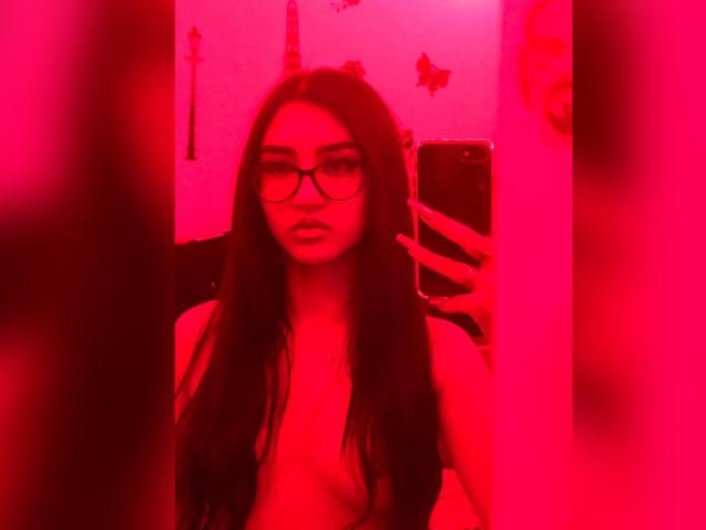 Selfie con illuminazione rossa di LuciaCalixto, con lunghi capelli scuri, occhiali e senza vestiti, che tiene un telefono, mostrando il seno, riflessa in uno specchio. I capezzoli non sono visibili..