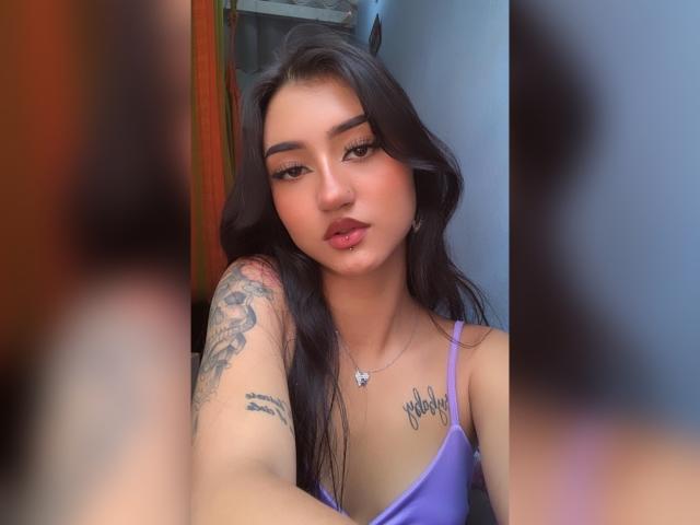 Lucia Calixto, avec ses longs cheveux noirs, son haut violet, son bras tatoué et ses lèvres pulpeuses, porte un collier avec un petit cœur, et montre sa poitrine ainsi qu'un léger bronzage..