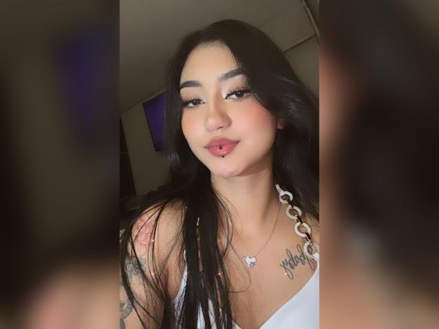 Lucia Calixto, con lunghi capelli neri, un anello rosa al labbro, una canotta bianca, un tatuaggio sulla spalla destra e una collana, sorride alla fotocamera, con le braccia sfocate in primo piano..