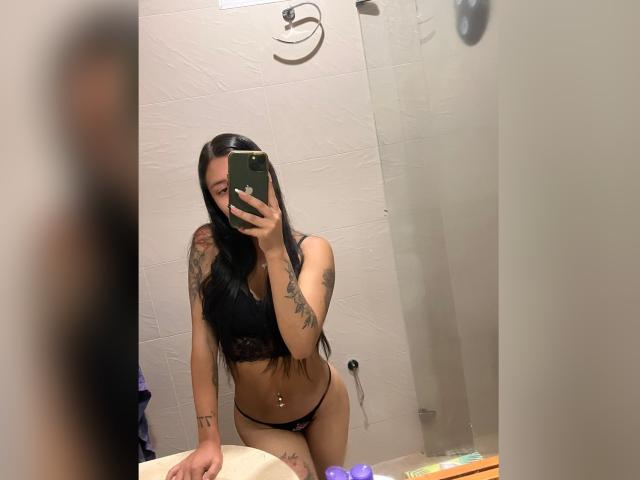 Lucia Calixto, com cabelos longos e pretos, vestindo um sutiã e calcinha de renda pretos, mostrando seus seios e vulva, com tatuagens no braço, segurando um celular, selfie no espelho no banheiro..
