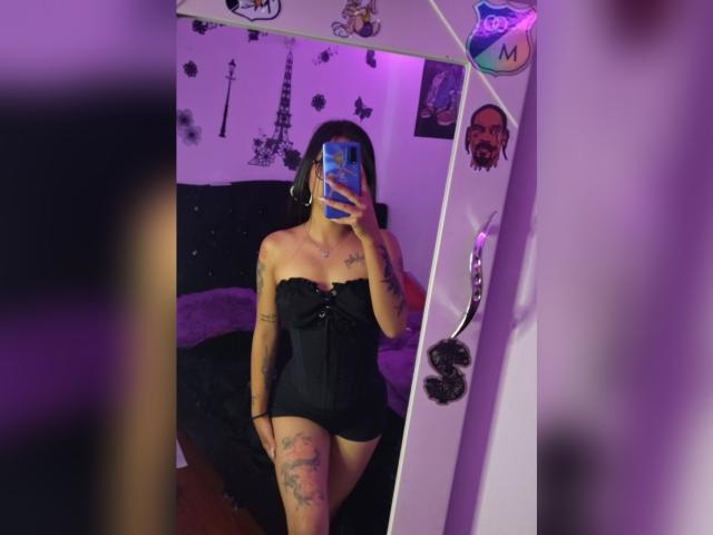 Lucia Calixto mit Tattoos, trägt ein schwarzes, trägerloses Korsett, das ihre Brüste und Vulva zeigt, und macht ein Selfie mit einem blauen Handy in einem violett beleuchteten Raum..