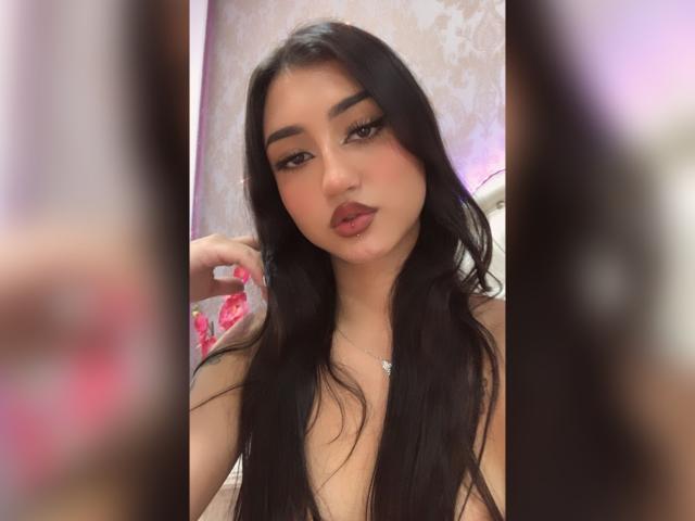 Lucia Calixto ha lunghi capelli neri, labbra rosse carnose e sopracciglia scure, guarda la fotocamera con un leggero sbuffo, con il seno visibile, indossando una collana d'argento..