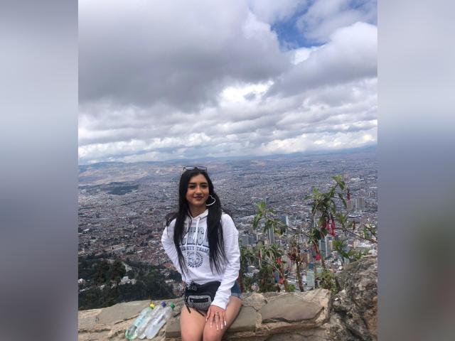 Lucia Calixto, met lang zwart haar, gekleed in een witte hoodie en korte broek, lachend, zittend op een stenen rand, met uitzicht op een uitgestrekte stad met een bewolkte lucht, met een zonnebril in haar hand..