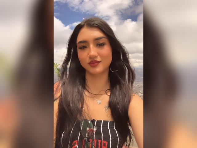 Lucia Calixto, com longos cabelos pretos, vestindo uma blusa preta sem alças, brincos grandes e batom vermelho, está em pé contra um céu azul nublado, mostrando uma tatuagem no lado direito do peito..
