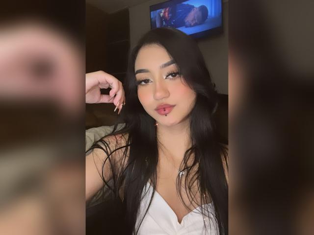 Lucia Calixto, com cabelos pretos longos, blusa branca e piercing rosa no lábio, segurando o queixo, olhando para a câmera, com um sorriso suave, olhos castanhos, seios médios, em um quarto com pouca luz..