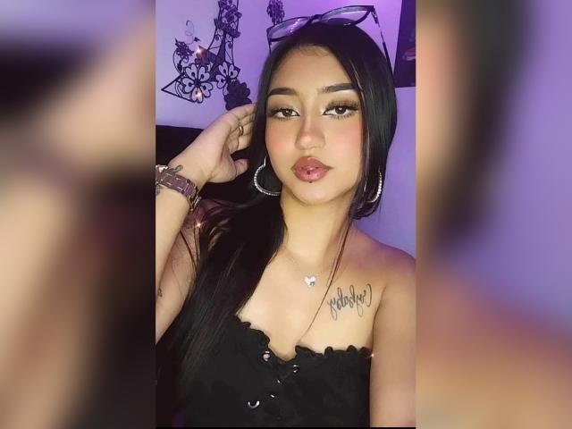LuciaCalixto with long black hair, wearing a black strapless top, showing her tits, a heart necklace, hoop earrings, and a tattoo on her right shoulder, with a serious expression.