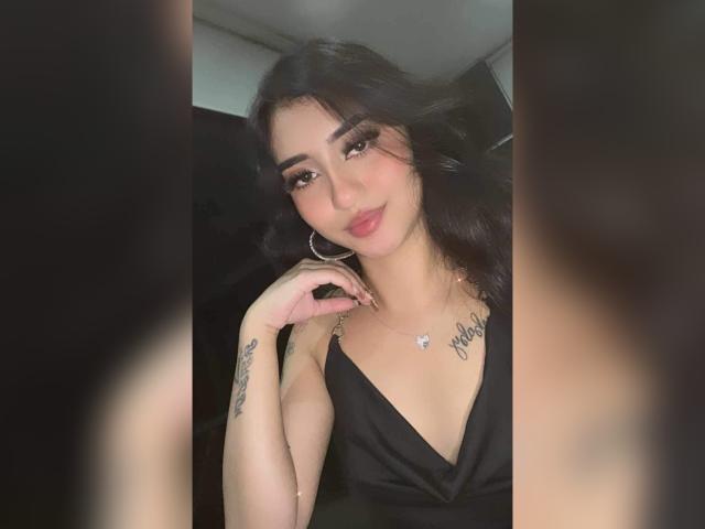 LuciaCalixto with black wavy hair, wearing a black dress, hoop earrings, and a necklace, showing tattoos on her right wrist, smiling with pink lipstick, and touching her chin.
