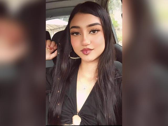 Lucia Calixto mit langem, schwarzem Haar, vollen Augenbrauen, großen, braunen Augen und pinkem Lippenstift, trägt ein schwarzes Oberteil, zieht die Lippen leicht zusammen und befindet sich in einem Auto..