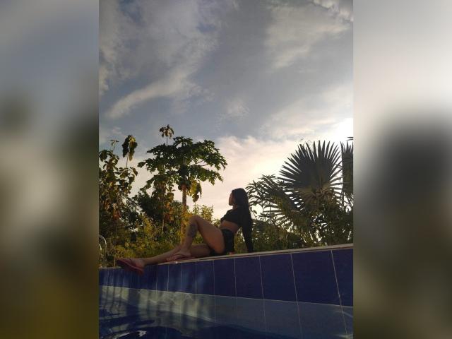 Lucia Calixto, assise sur le bord de la piscine en carreaux bleus, portant un maillot de bain noir, le soleil se couche, dos à la caméra, jambes croisées, palmiers et ciel en arrière-plan, tongs rouges à ses pieds..