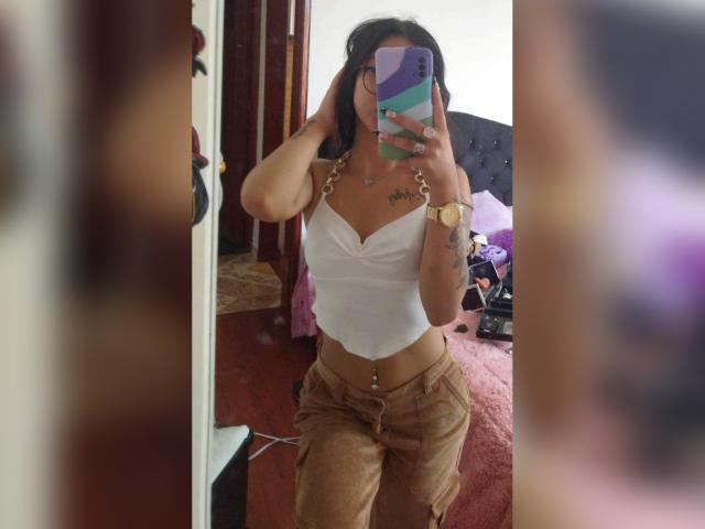 LuciaCalixto em uma selfie no espelho, vestindo uma cropped branca, calças bege, uma corrente dourada e um relógio, mostrando os seios e um piercing no umbigo, segurando um celular colorido..